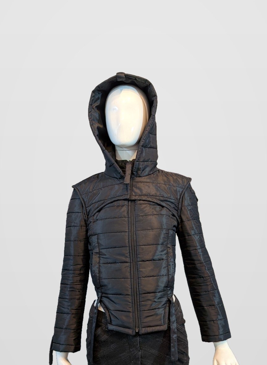 Black Nova Puffer — black futuristic quilted puffer jacket with hood on a mannequin, framed just below the jacket hem and side-strap ends, pearl-grey background
